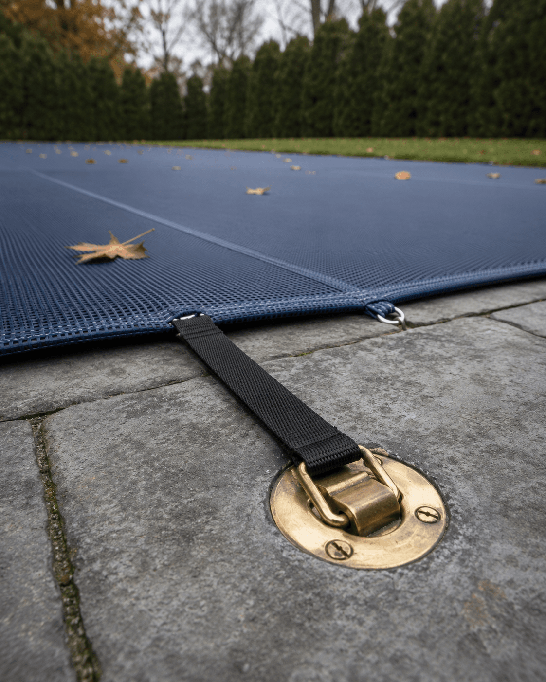 navy blue mesh pool safety cover brass anchor detail.png Close up of a navy blue mesh pool safety cover with a black tension strap and brass anchor mounted in grey paver decking, with scattered autumn leaves and a cedar hedge in the background.